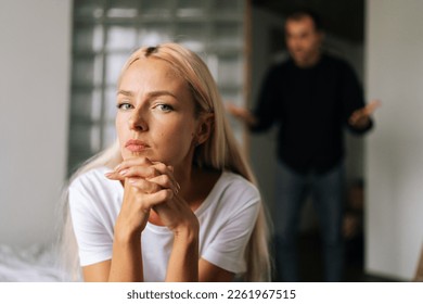 Portrait of tired young woman sadness looking at camera sitting on bed on background of aggressive husband shouting on girlfriend at home. Concept of family scandal, crisis, domestic violence, abuse.