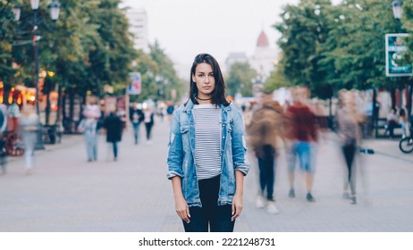retrato de una joven estudiante cansada parada sola en el centro de la ciudad mirando a la cámara con la cara recta mientras una multitud de hombres y mujeres se revolcan por ahí.