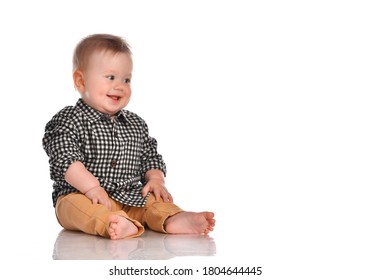 Portrait of a toddler baby boy with blue eyes wearing beige pants and a plaid shirt. The child is sitting barefoot and smiling happily on a white background with space for text.