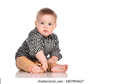 Portrait of a toddler boy with blue eyes in beige trousers and a plaid shirt. The child sits and looks at you on a white background with place for text.