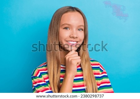Portrait of toothy beaming schoolgirl with long hairstyle wear colorful t-shirt smiling fingers on chin isolated on blue color background