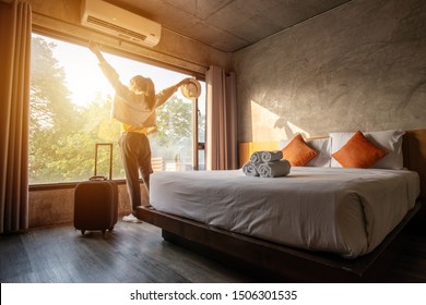 Portrait of tourist woman raised her hands and standing nearly window, looking to beautiful view with her luggage in hotel bedroom after check-in. Conceptual of travel and vacation.