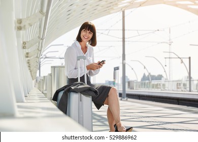 Portrait of traveling business woman with smart phone and luggage waiting on platform