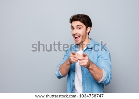 Portrait of trendy, brunet, manly, positive guy with stubble in shirt, having open mouth, pointing two forefingers front to the camera, isolated on grey background