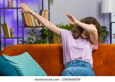 Portrait of trendy cheerful young woman having fun dancing and moving to rhythm, dabbing raising hands, making dub dance winner gesture celebrate sitting on sofa. Girl at home living room apartment