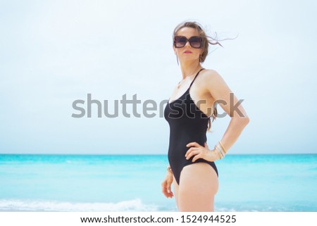 Portrait of trendy middle age woman with long curly hair in elegant black bathing suit on a white beach looking into the distance.