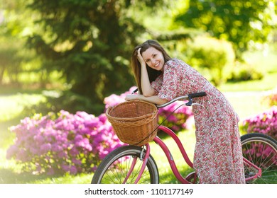 Portrait Of Trendy Young Woman In Long Pink Floral Dress Stop To Riding On Vintage Bike With Basket For Purchases, Flowers Background Outdoors. Pretty Female Recreation Time In Spring Or Summer Park