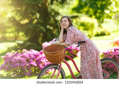 Portrait Of Trendy Young Woman In Long Pink Floral Dress Stop To Riding On Vintage Bike With Basket For Purchases, Flowers Background Outdoors. Pretty Female Recreation Time In Spring Or Summer Park