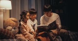 Portrait Of Two Active Children Engaged In A Conversation With Their Father While He Reads To Them Before Bed. Cute Korean Kids Not Ready To Go To Sleep, Reading A Story With Their Male Parent
