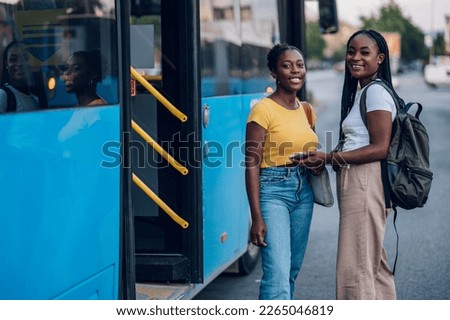 Portrait of a two african american woman waiting at a bus stop and talking cheerfully while looking into the camera. Diverse tourists in new town. Best friends traveling together in a city.