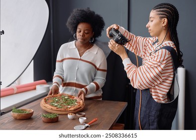 Portrait of two African American young women as female photographers working together during food photography set taking pictures of fresh rustic pizza on wooden table - Powered by Shutterstock