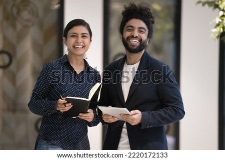 Portrait of two ambitious young 30s Indian male female colleagues pose in workspace smile look at camera, hold personal organizer and digital tablet. Professional occupation people, team-work, career