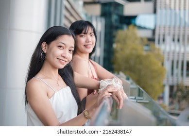 Portrait Of Two Asian Women Smiling And Hanging Out By The Ledge Of An Outdoor Mall.