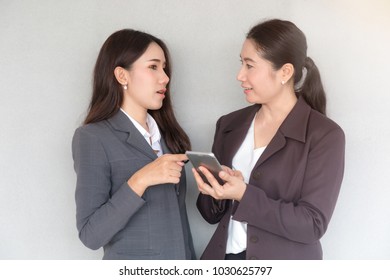 Portrait Of Two Attractive Young Asian Businesswoman Wearing A Suit Holding A Smartphone And Talking About Work Or New Technology With Eye Contact In The Office Background.