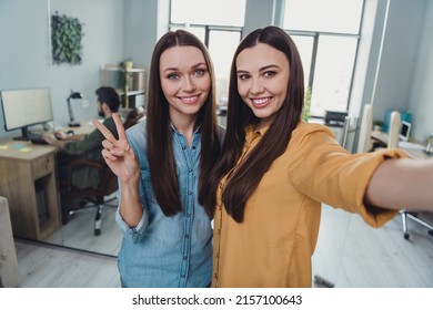 Portrait of two beautiful cheerful girls experts friends showing v-sign having fun good mood at workplace workstation indoors