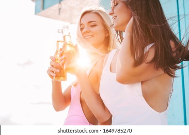 Portrait of two beautiful girls cheering and posing with bottles of beer at seaside. Young attractive women friends drinking cold beer and having fun on sunny beach. Summer travel and friendship
