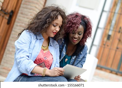 Portrait Of Two Beautiful Girls With Tablet Computer In Urban Background, Black And Mixed Women. Friends Talking