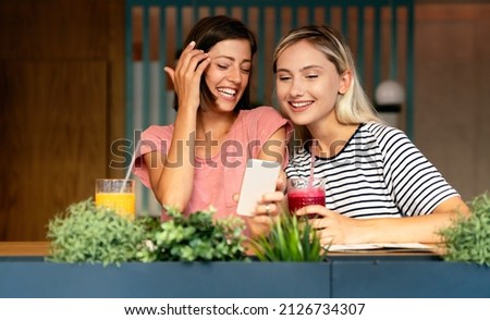 Portrait of two beautiful women having fun, using phone together and chatting in cafe