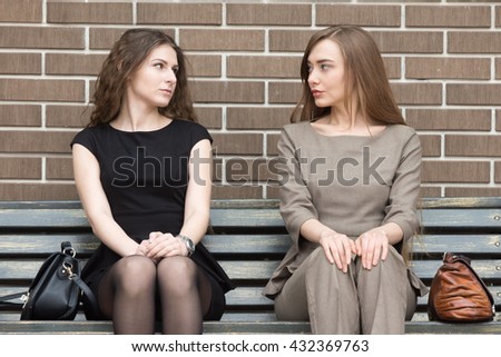 Portrait of two beautiful young female rivals sitting side by side on bench and looking at each other with challenging expressions. Attractive caucasian office women ready for confrontation