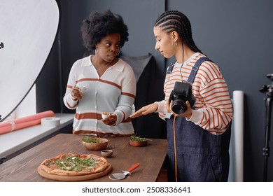 Portrait of two Black young women as team of female photographers collaborating during food photography set and staging fresh rustic pizza on wooden table - Powered by Shutterstock