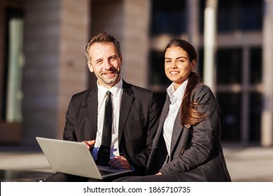 Portrait of two business peope, middle aged man and young woman in suits, smiling at camera while sitting on bench outdoors and discussing project on laptop