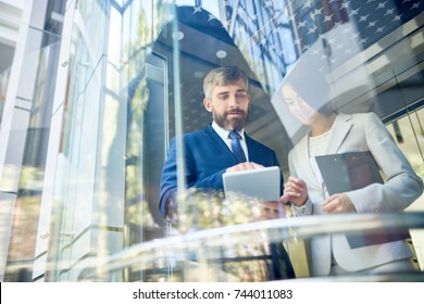 Portrait Of Two Business People, Man And Woman, Using Digital Tablet Discussing Work Standing Behind Window In Office Center, Copy Space