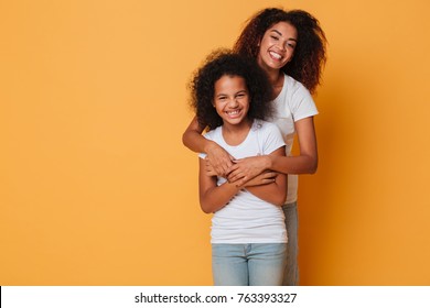 Portrait of two cheerful african sisters hugging and looking at camera isolated over orange background