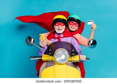 Portrait of two cheerful elderly retired pensioners riding moped using card buy order isolated over bright blue color background