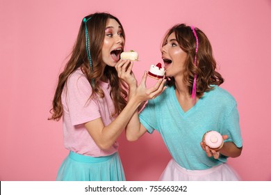 Portrait of two cheerful girls in colorful bright clothes eating cupcakes isolated over pink background