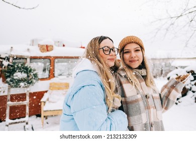 Portrait of two cheerful women friends standing outdoor near van during the snowfall in winter camp.