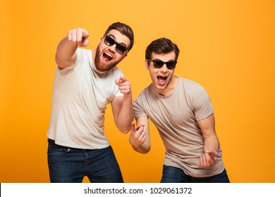 Portrait of a two cheerful young men in sunglasses pointing fingers at camera isolated over yellow background - Powered by Shutterstock