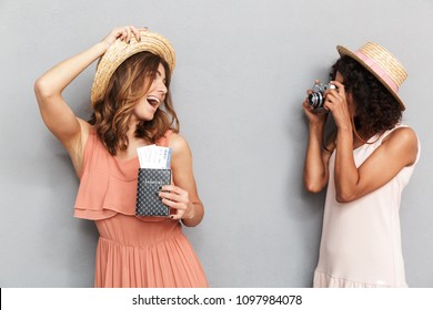 Portrait Of Two Cheerful Young Women Dressed In Summer Clothes Holding Passport With Tickets And Taking A Photo Over Gray Background