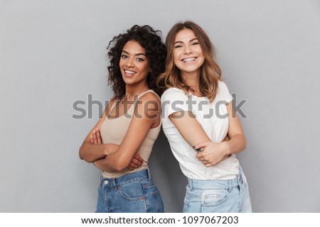 Portrait of two cheerful young women standing together and looking at camera isolated over gray background