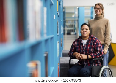 Portrait of two college students, one of them in wheelchair, posing together standing by shelves in library, copy space