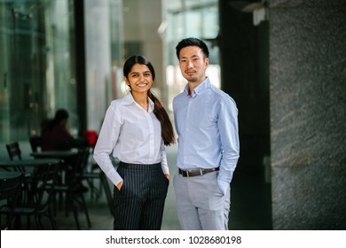 Portrait Of Two Coworkers, Team Mates Or Colleagues Standing In Their Office During Day. One Is An Indian Woman, And The Other A Chinese Man (diverse). They Are Smiling In A Relaxed Way. 