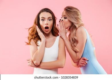 Portrait of a two cute girls dressed in swimsuits whispering a secret isolated over pink background