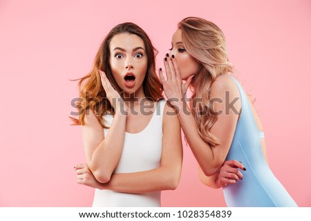 Portrait of a two cute girls dressed in swimsuits whispering a secret isolated over pink background