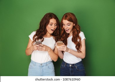 Portrait of two cute women with red hair using cell phones with surprise isolated over green background