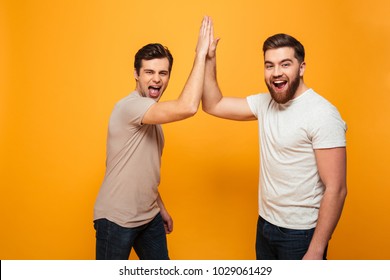 Portrait of a two delighted young men celebrating with high five gesture isolated over yellow background