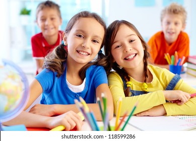 Portrait von zwei fleißigen Mädchen, die auf der Arbeitsfläche mit Schuljungen auf dem Hintergrund eine Kamera anschauen
