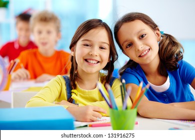 Portrait von zwei fleißigen Mädchen, die auf der Arbeitsfläche mit Schuljungen auf dem Hintergrund eine Kamera sehen