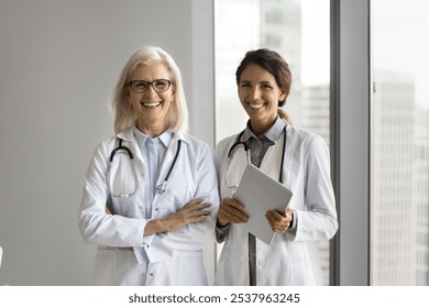 Portrait two diverse age female doctors pediatricians surgeons cardiologists standing in white coats. Medical specialists teammates modern healthcare center staff look at camera with confident smiles - Powered by Shutterstock