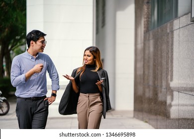 Portrait Of Two Diverse Asian Business People (colleagues Meeting For Lunch) Walking In The City (Singapore River). One Is A Korean Man, The Other A Malay Woman. They Are Both Smiling As They Chat.