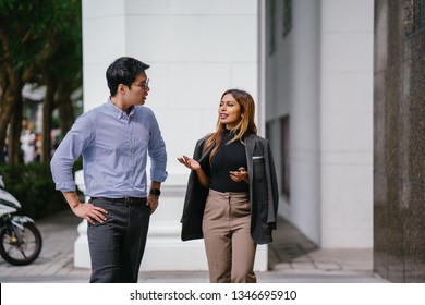 Portrait of two diverse Asian business people (colleagues meeting for lunch) walking in the city (Singapore River). One is a Korean man, the other a Malay woman. They are both smiling as they chat.