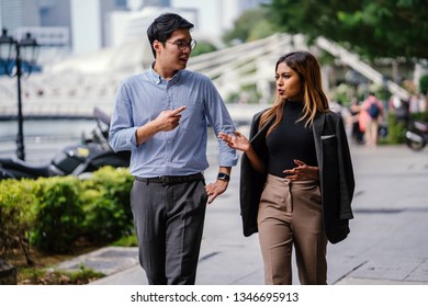 Portrait Of Two Diverse Asian Business People (colleagues Meeting For Lunch) Walking In The City (Singapore River). One Is A Korean Man, The Other A Malay Woman. They Are Both Smiling As They Chat.