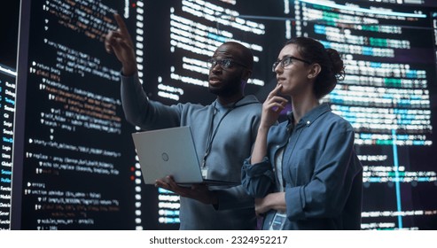 Portrait of Two Diverse Developers Using Laptop Computer, Discussing Lines of Code that Appear on Big Screens Surrounding Them. Male and Female Programmers Creating Software Together and Fixing Bugs - Powered by Shutterstock