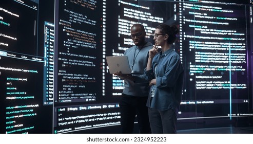 Portrait of Two Diverse Developers Using Laptop Computer, Discussing Lines of Code that Appear on Big Screens Surrounding Them. Male and Female Programmers Creating a Software Together, Fixing Bugs - Powered by Shutterstock