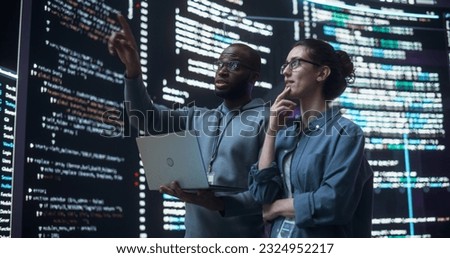 Portrait of Two Diverse Developers Using Laptop Computer, Discussing Lines of Code that Appear on Big Screens Surrounding Them. Male and Female Programmers Creating Software Together and Fixing Bugs