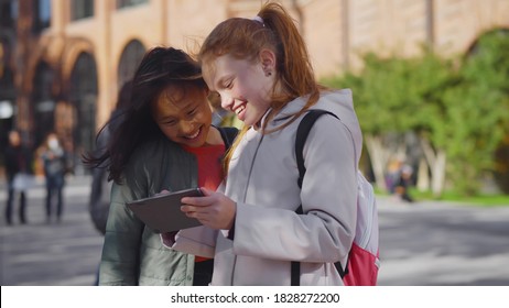 Portrait Of Two Diverse Schoolgirls Using Digital Tablet Standing Outdoors. Asian And Caucasian Girls Students Watching Photos On Tablet Pc Outside School Building After Classes