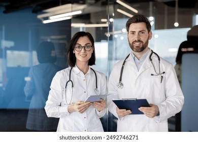 Portrait of two doctors standing together in hospital. Both wearing white coats and stethoscopes. Female doctor holding tablet while male doctor holding clipboard. - Powered by Shutterstock